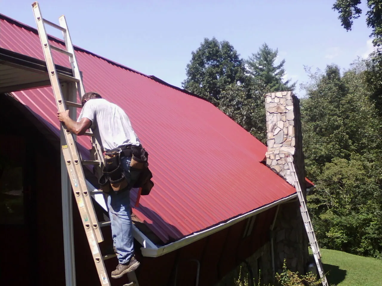 Roofer on a ladder beside a red metal roof and stone chimney — Soffit & Fascia Repair by Roofing Brothers in Red Bank